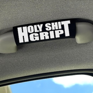 May include: A black car grab handle cover featuring the white text HOLY SHIT GRIP printed in a bold, sans-serif font. The cover is wrapped around a light grey interior vehicle ceiling handle, providing a contrasting visual element to the car interior.