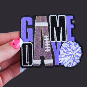 May include: A rectangular patch with the words "GAME DAY" in purple, black, and white. The letter "A" is designed to look like a football. A white and purple pom-pom is on the right side of the patch.