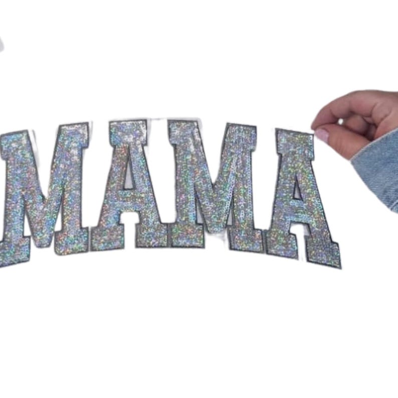 May include: Silver glitter "MAMA" applique with a collegiate font. The letters are outlined in a darker silver and are on a white background. A hand wearing a denim jacket is holding the applique.