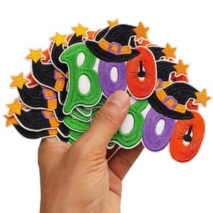 May include: A hand holding a collection of Halloween-themed patches. The patches feature the word "BOO" in green, purple, and orange, with white accents. Each patch has a witch hat with a gold star and orange accents.