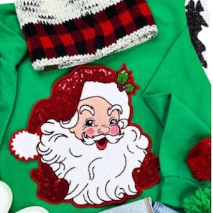 Puede incluir: Una camiseta verde de manga larga con un motivo de Papá Noel con lentejuelas. El diseño de Papá Noel presenta un gorro y guantes rojos con lentejuelas, una barba blanca y mejillas rosadas. En la parte superior se ve un gorro de punto a cuadros rojos y negros.