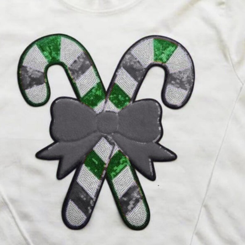 May include: White shirt with a sequined candy cane design. The candy canes are green and silver, with a gray bow at the center. The design is a festive, holiday-themed embellishment.