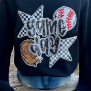 May include: Navy blue sweatshirt with the words "Game Day" in silver sequins. The design includes a baseball, a baseball glove, and two stars with a checkered pattern. The sweatshirt is worn with blue jeans.
