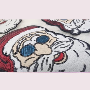 May include: Close-up of sequined Santa Claus face patches. The patches feature a red hat, white beard, and blue sequined eyes. The Santa face is outlined in black, with a beige face and red mouth.