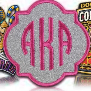 May include: An embroidered patch with the letters AKA in pink script. The patch has a silver glitter background and a pink border. Other patches are visible in the background.