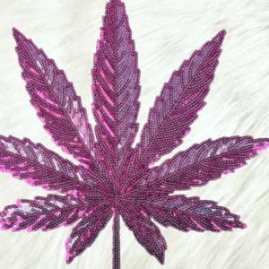 May include: A large, stylized purple cannabis leaf, heavily embellished with small, round beads. The leaf's veins are subtly highlighted with lighter pink beads, creating a textured, shimmering effect against a white, fluffy background.