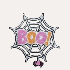May include: A Halloween patch with a black and white spiderweb design. The word "Boo!" is spelled out in pink and orange sequins. A pink spider hangs below the web, creating a festive and spooky decoration.