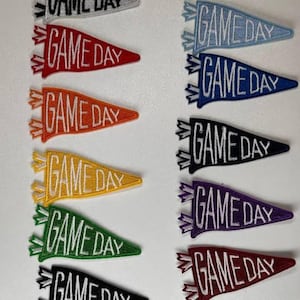 May include: A collection of pennant-shaped patches, each displaying the text "GAME DAY" in white. The patches are in a variety of colours: red, orange, yellow, green, blue, black, purple, and maroon. Each patch has a decorative border.