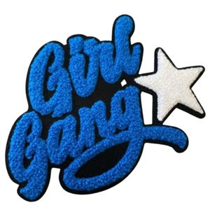 Op de afbeelding: Een patch met de woorden "Girl Gang" in blauwe, getextureerde letters. Een witte ster met een zwarte omtrek staat naast de tekst. De patch staat op een zwarte achtergrond.