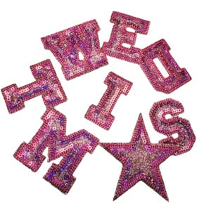 Puede incluir: Parches rosas con lentejuelas que deletrean la palabra "WE MISS" y una estrella. Los parches están en una variedad de fuentes y tamaños, con una mezcla de lentejuelas rosas y moradas. La estrella es brillante.