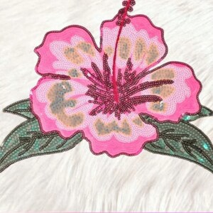 May include: A pink sequined hibiscus flower with a dark red center and a light brown pattern. The flower is surrounded by green leaves with black sequins. The design is on a white, textured background.