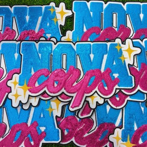 May include: A collection of patches with the word "NOVA" in blue sequins and "corps" in pink sequins. Each patch has a white border and yellow star accents. The patches are arranged on a green surface.