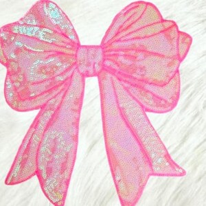 May include: A large, pink sequined bow with a shimmering, iridescent effect. The bow is a bright, eye-catching accessory, perfect for embellishing clothing, gifts, or crafts. The bow's design features multiple loops and long, flowing tails.