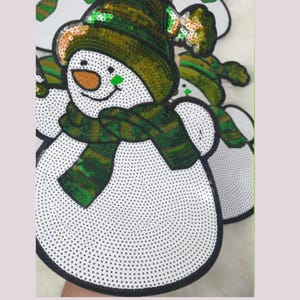 May include: White snowman applique with a black outline, adorned with a green and gold sequined hat and scarf. The snowman has a carrot nose and a green square on its cheek. The design is embellished with small black dots.