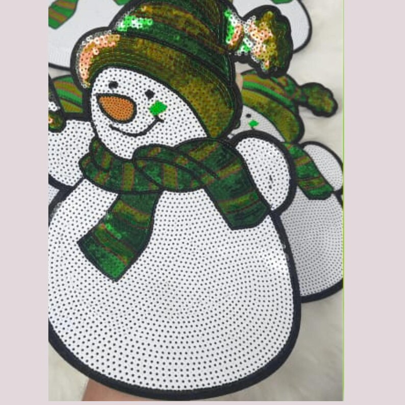 May include: White snowman applique with a black outline, adorned with a green and gold sequined hat and scarf. The snowman has a carrot nose and a green square on its cheek. The design is embellished with small black dots.