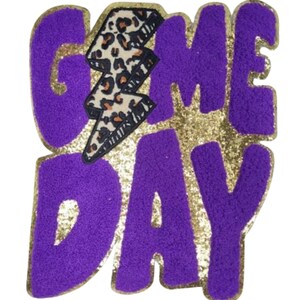 Könnte beinhalten: Ein lila-goldener Aufnäher mit den Worten "GAME DAY" in einer strukturierten Schriftart. Ein Blitz mit Leopardenmuster ist über dem Wort "DAY" zentriert. Der Aufnäher hat einen goldenen Glitzerhintergrund.