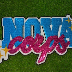 May include: A patch with the words "NOVA CORPS" in blue and pink sequins. The word "NOVA" is in a bold, blocky font, while "CORPS" is in a cursive script. Gold starbursts are on the left and right sides of the patch.