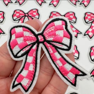 Peut inclure: Écusson brodé en forme de nœud papillon à carreaux roses et blancs, avec un contour noir et un dos blanc. Le nœud est tenu dans une main, avec plusieurs autres écussons similaires en arrière-plan.