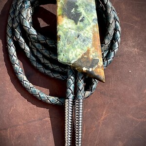 May include: A bolo tie featuring a rectangular, multi-colored stone pendant with shades of green, brown, and black. The pendant is attached to a braided, dark blue-grey leather cord, which is finished with two polished, silver-toned metal tips.