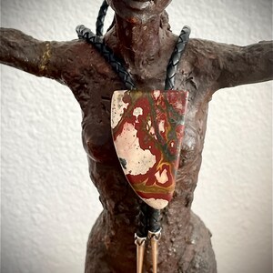 May include: A bolo tie featuring a large, triangular polished jasper stone with swirling patterns of deep red, cream, and olive green. The stone is suspended from a black braided leather cord, which is finished with two long, pointed silver-toned metal tips.