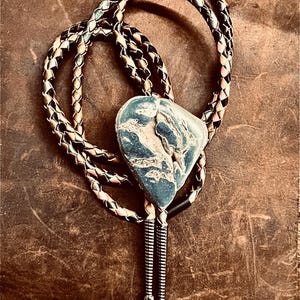 Op de afbeelding: Een bolo stropdas met een blauw-witte stenen hanger. De hanger hangt aan een gevlochten koord met zwarte, witte en roze accenten. De stropdas heeft een zilverkleurige metalen sluiting.