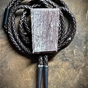 May include: A bolo tie featuring a rectangular, textured pendant with a brown and white pattern. The pendant is suspended from a braided brown leather cord with silver-toned tips. The overall design is rustic and stylish.