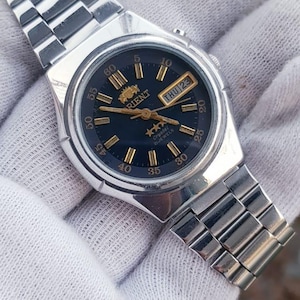 May include: Silver Orient watch with a dark blue face, gold hands, and markers. The watch shows the day and date. The watch band is silver metal. The watch is held in a white glove. The watch face has a diameter of approximately 3.5 cm.