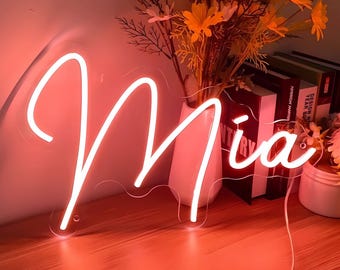 May include: A pink neon sign with the name "Mia" in a cursive font. The sign is lit up and casting a warm glow on the wooden surface it is sitting on.
