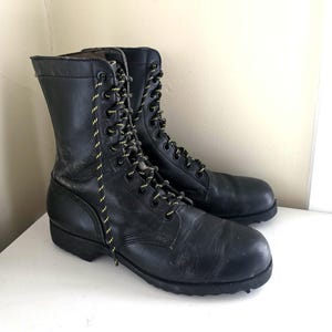 May include: A pair of black leather combat boots featuring a high-top design, rounded toes, and thick black rubber soles. The boots are secured with black laces that have thin yellow diagonal stripes, threaded through black metal eyelets.