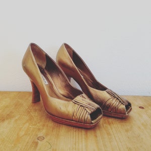 May include: A pair of gold peep-toe heels with a high heel and a platform sole. The shoes are made of leather and have a gold metallic finish.