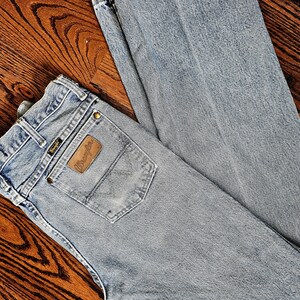 May include: Light blue Wrangler jeans with a brown leather patch on the back pocket. The jeans are laid out on a wooden surface. The Wrangler logo is visible on the patch.