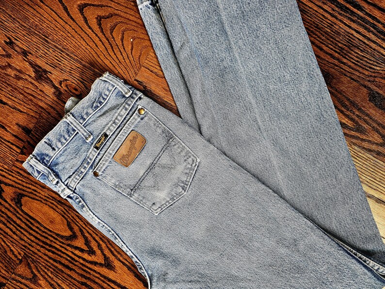 May include: Light blue Wrangler jeans with a brown leather patch on the back pocket. The jeans are laid out on a wooden surface. The Wrangler logo is visible on the patch.