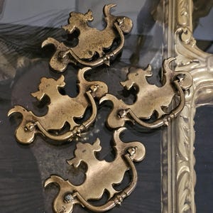 May include: Four antique brass drawer pulls with a decorative, stylized design. Each pull features a curved handle and a detailed, ornate shape, likely for a cabinet or furniture restoration project. The pulls are mounted on a dark surface.