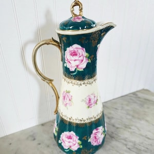 May include: A tall, green and white ceramic pitcher with a gold trim and pink rose floral pattern. The pitcher has a lid with a gold knob.