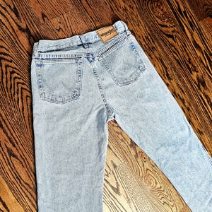 May include: Light blue denim jeans with a classic five-pocket design. The jeans feature a Wrangler brand patch on the back waistband. The jeans are laid flat on a wooden surface.