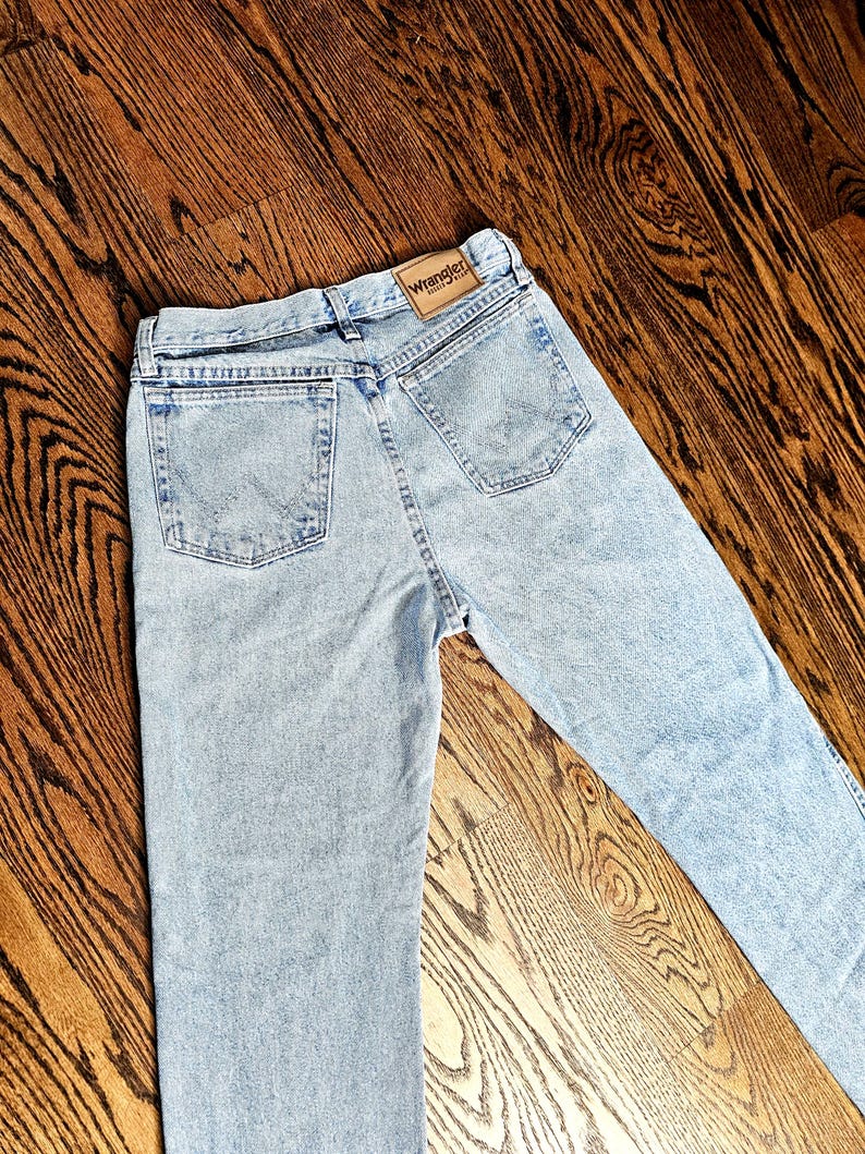 May include: Light blue denim jeans with a classic five-pocket design. The jeans feature a Wrangler brand patch on the back waistband. The jeans are laid flat on a wooden surface.