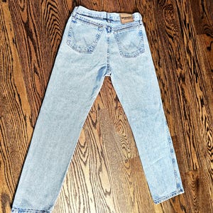 May include: Light blue Wrangler jeans with a classic five-pocket design. The jeans have a straight leg cut and a faded wash. The Wrangler logo is visible on the back pocket. The jeans are laid out on a wooden floor.