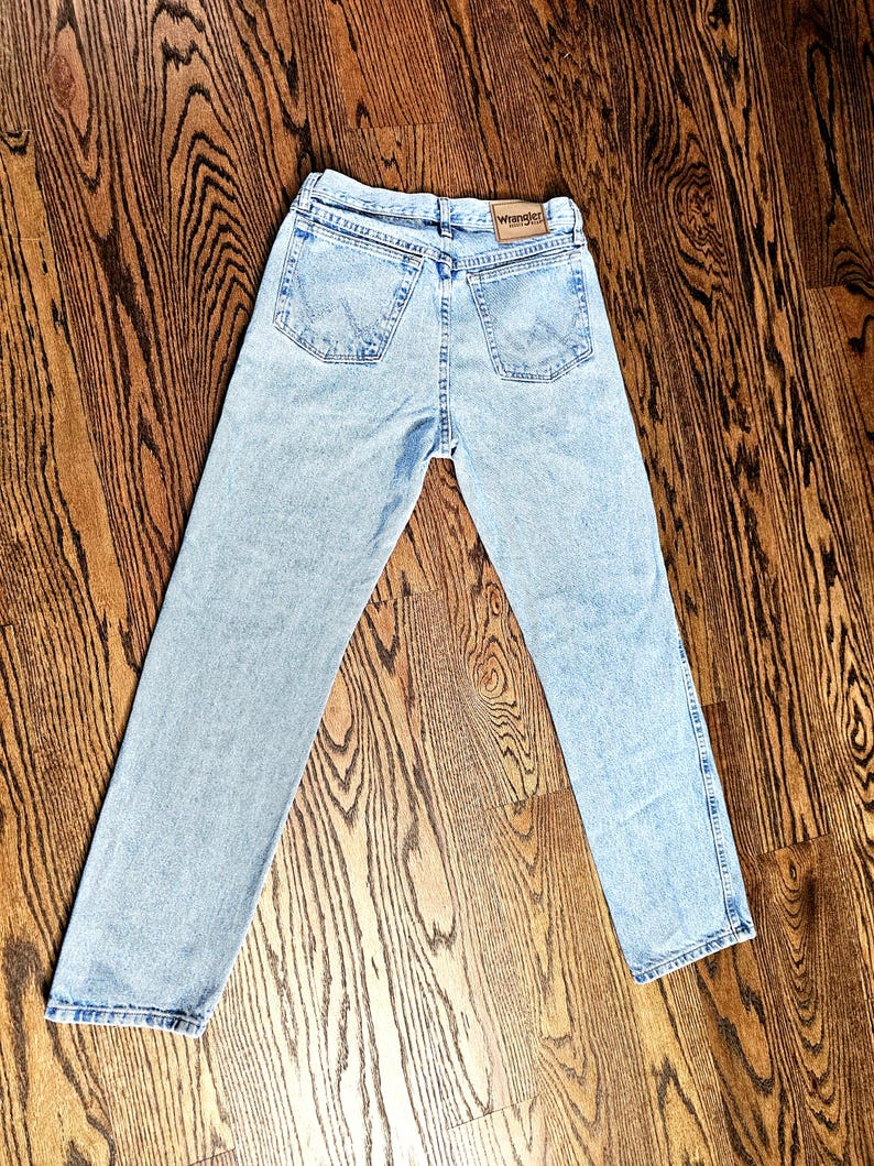 May include: Light blue Wrangler jeans with a classic five-pocket design. The jeans have a straight leg cut and a faded wash. The Wrangler logo is visible on the back pocket. The jeans are laid out on a wooden floor.