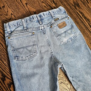 May include: Light blue denim jeans with classic five-pocket styling. Features include a brown leather patch with the brand name and a stitched design on the back pockets. The jeans are displayed on a wooden surface.