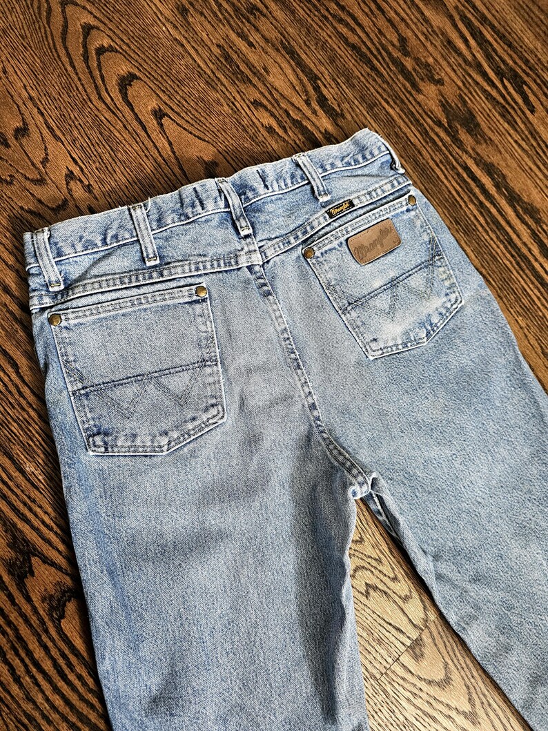 May include: Light blue denim jeans with classic five-pocket styling. Features include a brown leather patch with the brand name and a stitched design on the back pockets. The jeans are displayed on a wooden surface.