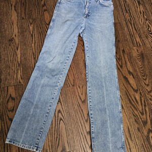 May include: Light blue denim jeans with a classic five-pocket design. The jeans have a straight leg cut and a button and zipper closure. The denim has a faded wash, giving them a vintage look. The jeans are laid flat on a wooden surface.