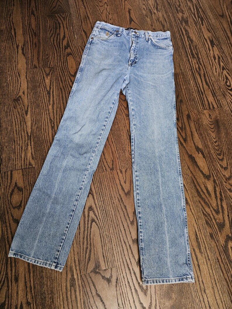 May include: Light blue denim jeans with a classic five-pocket design. The jeans have a straight leg cut and a button and zipper closure. The denim has a faded wash, giving them a vintage look. The jeans are laid flat on a wooden surface.