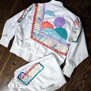 May include: White track suit with colorful geometric and abstract patches. The jacket has a high collar and ribbed cuffs. The pants are loose-fitting. The patches include red, orange, teal, and purple accents. The suit is made of a lightweight material.