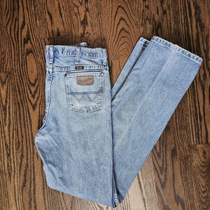 May include: Light blue Wrangler jeans with a classic five-pocket design. The jeans feature a brown leather patch on the back pocket with the Wrangler logo. The denim pants are laid out on a wooden surface.