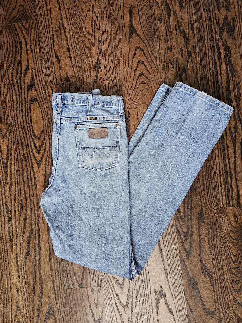 May include: Light blue Wrangler jeans with a classic five-pocket design. The jeans feature a brown leather patch on the back pocket with the Wrangler logo. The denim pants are laid out on a wooden surface.