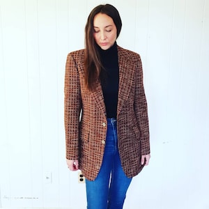 May include: A brown tweed blazer with a subtle plaid pattern. The blazer has gold-colored buttons and is worn with a black turtleneck and blue jeans. The jacket is a classic style.