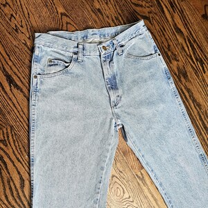 May include: Light blue denim jeans with classic five-pocket styling. The jeans have a button and zipper closure, belt loops, and a faded wash. The jeans are laid flat on a wooden surface.