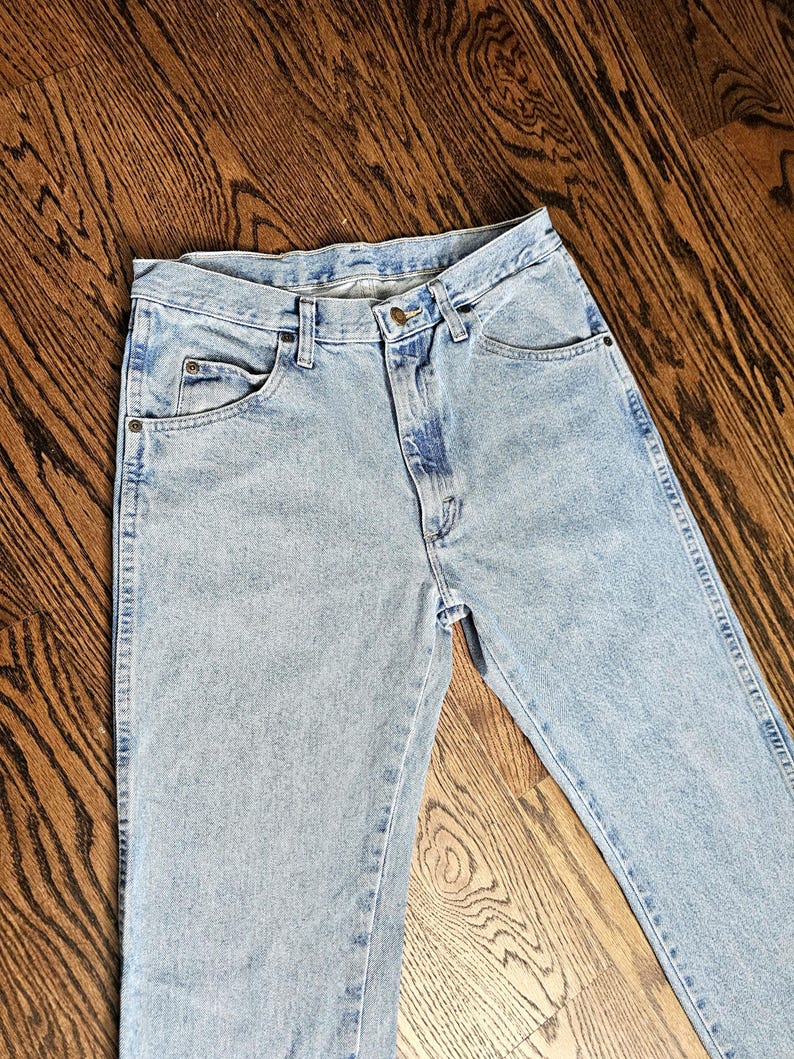 May include: Light blue denim jeans with classic five-pocket styling. The jeans have a button and zipper closure, belt loops, and a faded wash. The jeans are laid flat on a wooden surface.