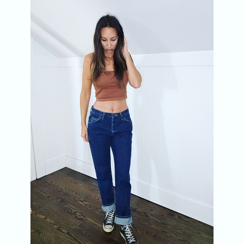 May include: A woman wearing a brown crop top and dark blue jeans with the cuffs rolled up. She is wearing black canvas sneakers.