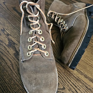 May include: Brown suede ankle boots with tan laces and brass eyelets. The boots have a rounded toe and a thick black sole. The boots are displayed on a wooden surface.